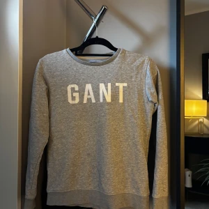 Grå sweatshirt från GANT - Snygg grå sweatshirt från GANT med vit logga framtill. Klassisk rund halsringning och långa ärmar. Perfekt för en avslappnad och stilren look. Tillverkad i mjuk bomullsmix som känns skön mot huden.