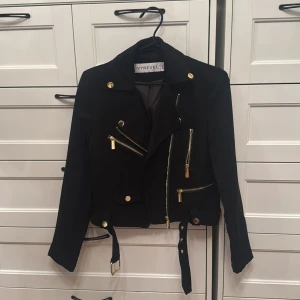 Svart mocka bikerjacka från Ivyrevel - Snygg svart bikerjacka från Ivyrevel i mockaliknande material med guldiga detaljer, flera dragkedjor och bälte i midjan. Jackan är croppad och har långa ärmar samt coola knappar på kragen och vid ärmslut. Som ny!