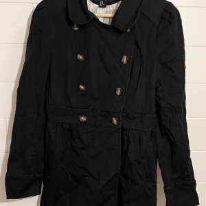 Svart trenchcoat med knappar - Stilren svart trenchcoat från H&M med dubbelknäppning och klassisk krage. Jackan har markerad midja, långa ärmar och dekorativa knappar framtill. Perfekt för dig som gillar en tidlös och snygg look.