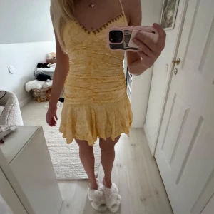 Gul kort klänning med volangkant - Superfin gul kort klänning med smala axelband och v-ringning dekorerad med små detaljer. Klänningen har rynkade partier över hela livet och en volangig kjol med ojämn kant. Tunt, lätt material med diskret blommönster och somrig vibe.