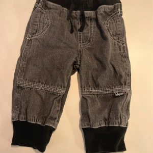 Grå jeansbyxor med svarta muddar Baby Star - Grå jeansbyxor från Baby Star med svarta muddar i midja och benslut. Byxorna har snörning i midjan, vita kontrastsömmar och två framfickor. Perfekta för en chill och bekväm stil till de minsta.