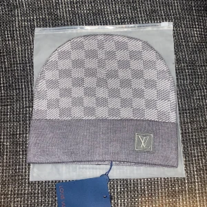 Grå rutig mössa från Louis Vuitton - Snygg grå mössa från Louis Vuitton med klassiskt rutigt mönster och uppvikt kant. Mössan har en diskret LV-logga framtill och är tillverkad i mjukt material som känns lyxigt. Perfekt accessoar för att lyfta din vinterstil.