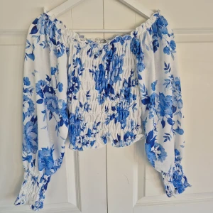 Blommig offshoulder blus i blått och vitt - Superfin offshoulder blus med blått blommönster på vit botten. Toppen har långa puffiga ärmar och smockad kropp. Storlek S som endast är använd vid ett tillfälle, nyskick!