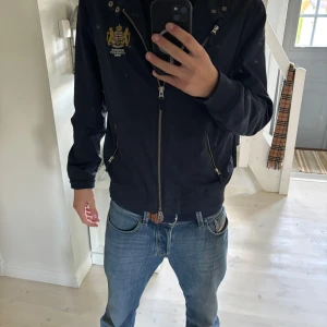 Morris Company marinblå bomberjacka - Snygg marinblå bomberjacka från Morris Company med guldbroderad logga på bröstet och Union Jack-flagga på insidan. Jackan har flera dragkedjor, ribbade muddar och klassisk krage. Perfekt för dig som gillar stilren design med brittisk touch.