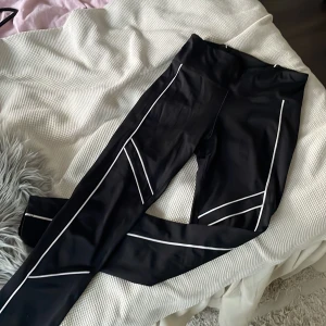 Svarta leggings från Stronger med vita detaljer - Svarta leggings från Stronger med vita linjer längs benen som ger en sportig vibe. Hög midja och tight passform, perfekta för träning. Tillverkade i ett stretchigt syntetmaterial som sitter skönt och följer kroppen. ✨