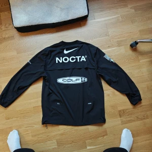Svart NOCTA x Nike långärmad tröja - Svart långärmad tröja från Nike NOCTA med stora vita tryck på ryggen och ärmarna. Crewneck, elastiska muddar och dragsko nertill. Snygg streetwear-vibe och perfekt för golf eller chill dagar. Materialet är lätt och känns som syntet.