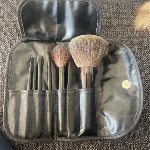 MAC sminkborstar i svart fodral - Snyggt set med fem sminkborstar från MAC i ett svart, stilrent fodral i konstläder. Innehåller borstar för både ansikte och ögon, perfekt för att fixa hela sminklooken. Borstarna har svarta handtag och mjuka borst i naturfärgade toner.