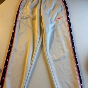 Nike repeat byxor - Snygga vita mjukisbyxor från Nike med elastisk midja och svart sidopaneler med vita Nike-loggor och rosa kant. Byxorna har fickor framtill och en rosa Nike Swoosh på ena benet. Perfekta för chill eller träning. storlek S men passar för M