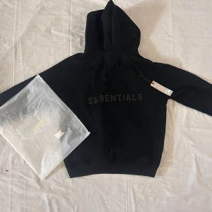Svart Essentials Fear of God hoodie L - Svart hoodie från Essentials Fear of God i storlek L. Tröjan har stor huva, tryckt logga på bröstet och ryggen samt ribbade muddar. Tillverkad i 100% bomull för en skön och avslappnad vibe. Perfekt för dig som gillar streetwear och minimalistisk stil.