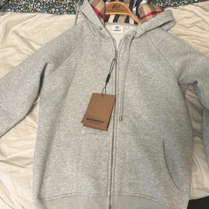 Grå hoodie från Burberry med dragkedja - Snygg ljusgrå hoodie från Burberry med klassiskt rutigt foder i huvan. Tröjan har dragkedja, snörning i huvan och broderad logga på bröstet. Perfekt för dig som gillar stilrena och exklusiva plagg med streetkänsla. Är helt sprillans ny och är för liten för mig säljer den för 700 pris kan diskuteras 
