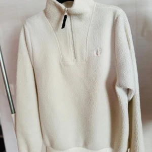 Vit fleece-tröja med half zip - Mysig vit fleece-tröja med hög krage och half zip-dragkedja. Tröjan har lång ärm och en enkel, clean look med diskret broderad logga på bröstet. Perfekt för kyliga dagar när du vill vara både bekväm och stilren.