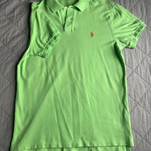 Grön pikétröja från Ralph Lauren - Snygg ljusgrön pikétröja från Polo Ralph Lauren i bomull. Klassisk krage, korta ärmar och röd broderad logga på bröstet. Custom fit-modell med knappar vid halsen och små slitsar i sidorna.