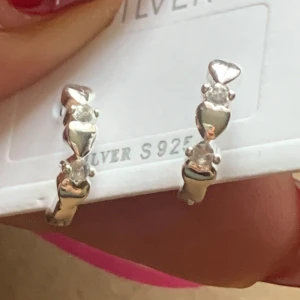 S925 silverörhängen med hjärtan - Snygga örhängen i äkta S925 silver med små hjärtan och gnistrande stenar. Designen är modern och söt, perfekt för dig som gillar detaljer. Hjärtana och stenarna sitter omlott längs hela örhänget och ger en glansig look.