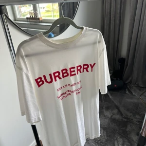 Vit Burberry t-shirt med röd logga - Vit t-shirt från Burberry med stor röd logga och text framtill. Storlek Small i nyskick.