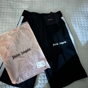 Palm Angels svarta shorts med vita ränder - Svarta shorts från Palm Angels med vita sidoränder och broderad logga framtill. De har elastisk midja och två fickor. Materialet är en mix av bomull och polyester, vilket ger en sportig och skön känsla. Dem är helt oanvända bara testade taget är fortfarande kvar 