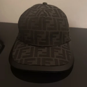 Svart Fendi keps med FF-mönster - Snygg svart keps från Fendi med det ikoniska FF-logomönstret över hela kepsen. Justerbar baktill med metallspänne där Fendi Roma är ingraverat. Kepsen är tillverkad i polyester och har en klassisk böjd skärm. Perfekt för dig som gillar exklusiva streetwear-accessoarer.