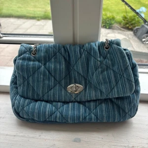 Blå quiltad axelväska från Becksöndergaard - Snygg axelväska från Becksöndergaard i blått randigt bomullstyg med quiltad design. Väskan har silverfärgad kedjerem och vridlås i metall. 