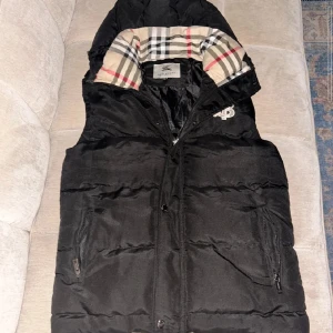Svart dunväst från Burberry - Svart dunväst från Burberry med klassiskt rutigt foder i huvan och broderad TB-logga på bröstet. Västen har dragkedja, knappar och två fickor med dragkedja. Perfekt för lager-på-lager och snygg streetstil. Pris kan diskuteras och byten går, skriv om ni har frågor