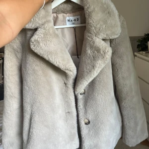 Beige/grå pälsjacka från NA-KD, stl 36 - Supermjuk och fluffig beige pälsjacka från NA-KD i storlek 36. Jackan har bred krage, knappar framtill och är i en kortare modell. Perfekt för dig som vill ha en mysig och trendig look under kyliga dagar.