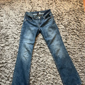 Blå low waist bootcut jeans från Videdress, stl 38 - Snygga blå bootcut jeans från Videdress i storlek 38. Klassisk femficksmodell med coola bakfickor med lock och knappar. Jeansen har en normal passform och är tillverkade i jeansmaterial med lätt slitning framtill för en trendig look.