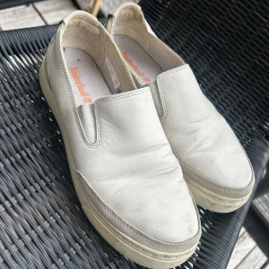 Beige slip-on skor från Timberland - Snygga slip-on skor från Timberland i beige skinn med diskret logga bak och elastiska paneler vid sidorna. Skorna har en platt sula och rund tå, samt detaljer i mocka vid hälen och tån. Perfekta för en chill och stilren look.