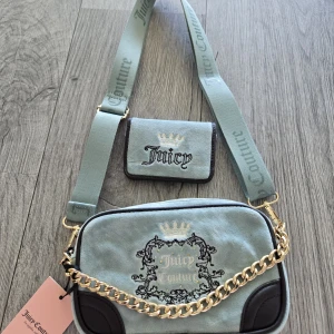 Juicy Couture axelväska med plånbok - Ljusblå axelväska från Juicy Couture i mjuk sammet med broderad logga och guldfärgad kedja. Matchande liten plånbok ingår. Väskan har bred axelrem med tryckt logga och svarta detaljer i hörnen. Insidan är fodrad med mönstrat tyg med Juicy Couture-tryck.