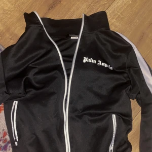 Palm Angels svart zip hoodie M - Svart hoodie från Palm Angels med vit dragkedja och vita detaljer längs ärmarna. Klassisk logga på bröstet och två fickor med dragkedja. Sportig och trendig modell i syntetmaterial, perfekt för en streetwear-look.