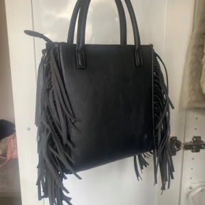 Svart handväska med fransar i skinn - Cool svart handväska i skinn med långa fransar på sidorna. Väskan har två handtag och en stilren, fyrkantig form. Perfekt accessoar för dig som vill sticka ut med en edgy och trendig look.