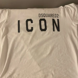 Vit Dsquared2 ICON t-shirt - Vit t-shirt från Dsquared2 med stor svart 'ICON'-text och logga tryckt på bröstet. Klassisk passform med rund hals och korta ärmar. Tillverkad i mjuk bomull som känns skön mot huden. Perfekt för dig som gillar streetstyle och vill sticka ut.