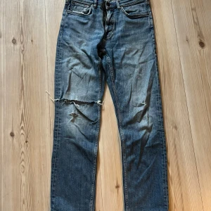 Blå jeans från Gant med slitningar 31/32 - Säljer ett par blå jeans från Gant i regular fit med klassisk femficksdesign. Jeansen har tydliga slitningar och hål på ena benet för en cool, avslappnad look. Tillverkade i bomull med skön passform och snygg tvättad finish.