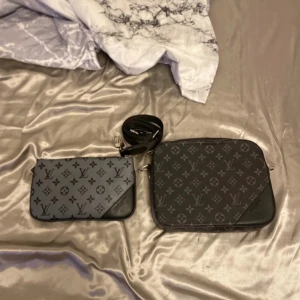 svart axelväska pris kan diskuteras  - Säljer en svart axelväska med klassiskt monogrammönster i grått. Väskan har två delar, båda med dragkedja och avtagbar axelrem. Tillverkad i skinn med detaljer i svart och grått. Perfekt för dig som vill ha en stilren och lyxig accessoar. Kedjan är lite konstig men går fortfarande att öppna och stänga 