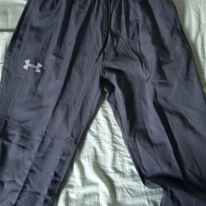 Svarta träningsbyxor från Under Armour - Svarta träningsbyxor från Under Armour med dragsko i midjan och dragkedja vid benslut. Byxorna har en sportig look med logga på låret och reflekterande detaljer. Perfekta för gymmet eller löprundan. Står storlek L men passar som S