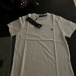 Vit t-shirt från Polo Ralph Lauren - Vit t-shirt från Polo Ralph Lauren med klassisk rund hals och korta ärmar. På bröstet finns den ikoniska broderade loggan i svart och en grön tryckt ruta med texten 'Polo by Ralph Lauren'. Skön passform och tillverkad i mjuk bomull.