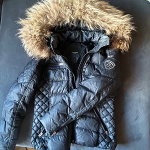 Svart pufferjacka Rockandblue päls - Svart pufferjacka från Rockandblue med stor fluffig päls på huvan. Jackan har quiltade detaljer på sidorna och ärmarna, dragkedjor på fickor och ärmar samt Rockandblue-logga på ena ärmen. Ett litet hål som säkert kan sys. Lite sminkfläckar men inte så illa