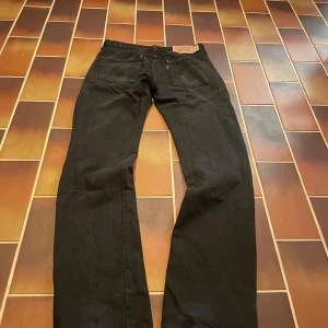 Svarta Levi's 501 jeans straight fit - Svarta Levi's 501 jeans med klassisk rak passform och femficksdesign. Jeansen är tillverkade i robust denim och har knäppning framtill. Snyggt slitna detaljer nertill på benen för en cool look. Perfekta för dig som gillar tidlös stil.