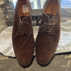 Bruna mockaskor från Sebago, stl 45 - Snygga bruna mockaskor från Sebago med klassisk design och rund tå. Skorna har snörning och en stilren siluett som passar perfekt till både jeans och kostym. Tillverkade i mjuk mocka med läderfoder för extra komfort. Levereras med originalpåse och kartong.Ordinarie  pris 3000kr