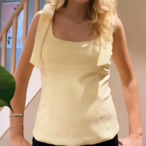 Gul linnetopp med rosett - Söt ljusgul linnetopp med breda axelband och en stor rosett på axlarna. Toppen har rak passform och fyrkantig ringning, perfekt för varma dagar. 