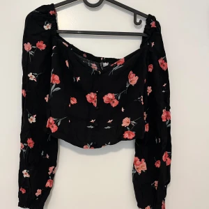 Svart blommig offshoulder blus - Supersöt svart blus från Divided med rosa och röda blommor. Offshoulder-modell med långa puffiga ärmar och knappar framtill. Perfekt för dig som gillar romantiska vibes och vill sticka ut lite extra.