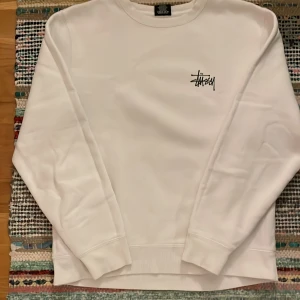 Vit Stüssy sweatshirt med logga - Vit crewneck sweatshirt från Stüssy med svart logga tryckt på bröstet och en stor logga på ryggen. Tröjan har rund hals. 