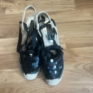 Svarta espadriller med paljetter Replay - Snygga espadriller från Replay med svart tyg, dekorerade med svarta paljetter och knytband runt vristen. Skorna har en hög flätad platåsula i beige och insida med metallic-detaljer. Perfekta för att lyfta din sommarstil.