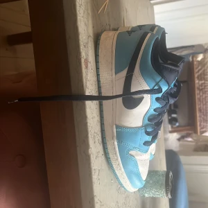 Nike Air Jordan 1 Low UNC blå/vit - Säljer ett par Nike Air Jordan 1 Low i UNC färgway med ljusblå och vita paneler, svart swoosh och mörkblå snörning. Skorna har rund tå, platt sula och klassisk Jordan-logga broderad på hälen. Materialet är skinn och de har perforeringar på tån för extra ventilation.