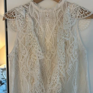Vit spetsblus med volangdetaljer - Superfin vit blus i spets med vackra volangdetaljer och hög hals. Blusen är ärmlös och har ett intrikat spetsmönster som ger en romantisk och bohemisk känsla. Perfekt för dig som vill sticka ut med en unik topp.