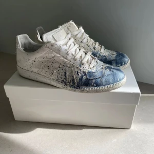 maison margiela  - Unika sneakers i vitt tyg med blå färg och coola färgstänk över hela skon. Lågt skaft, snörning och platt sula. Tåpartiet har en tydlig blå toning som ger en kreativ och edgy vibe. Perfekta för dig som vill sticka ut med din stil.
