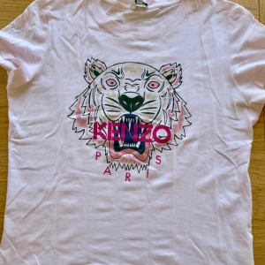 Ljusrosa Kenzo t-shirt med tigertryck - Kenzo t-shirt i ljusrosa med ikoniskt tigertryck och logga i rosa och blått framtill. Klassisk rund hals och korta ärmar. Tillverkad i mjuk bomull, perfekt för en cool och avslappnad stil.