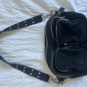 Svart axelväska från Gina Tricot - Cool svart axelväska från Gina Tricot med bred axelrem dekorerad med silvriga nitar. Väskan har flera fack med dragkedjor och två stora fickor framtill. Perfekt för dig som vill ha en praktisk men snygg väska med lite edge.