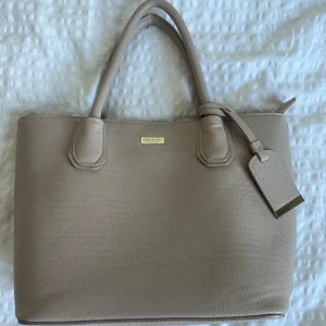 Beige handväska från Don Donna - Stilren beige handväska från Don Donna i skinnimitation med både handtag och avtagbar axelrem. Väskan har guldfärgade detaljer, dragkedja upptill och flera fack inuti för smart organisering. Perfekt storlek för att få plats med allt du behöver.