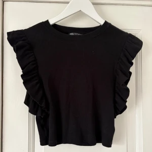 Svart volangtopp från Zara - Svart topp från Zara med volangdetaljer längs axlarna. Modellen är ärmlös och har en rund halsringning. Toppens material är mjuk bomull.
