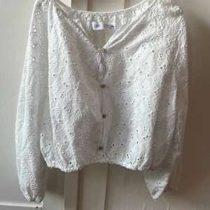 Vit broderad blus från Zara - Superfin vit blus från Zara med broderade blommönster och snygga träknappar framtill. Blusen har långa ärmar med resår vid ärmslut och en lätt croppad passform. Perfekt för dig som gillar romantiska detaljer och en luftig känsla.