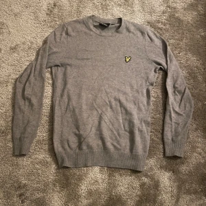 Grå tröja från Lyle & Scott - Säljer en stilren grå tröja från Lyle & Scott med deras klassiska gula fågel-logga på bröstet. Tröjan har rund halsringning, långa ärmar och ribbade muddar vid ärmslut och nederkant. Perfekt för dig som gillar enkel och snygg stil.