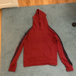 Röd hoodie från GANT med logga - Röd hoodie från GANT med huva och känguruficka. Svarta band med GANT-logga längs ärmarna ger en sportig vibe. Mjuk bomullskänsla och ribbade muddar. Perfekt för chill dagar eller när du vill ha en snygg streetstyle-look. Passar bäst för 170cm 15 år
